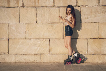 Young woman inline skates using smartphone leaning on wall - DLTSF00892