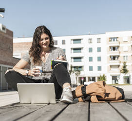 Smiling woman holding drink writing in book while sitting on boardwalk during sunny day - UUF20749