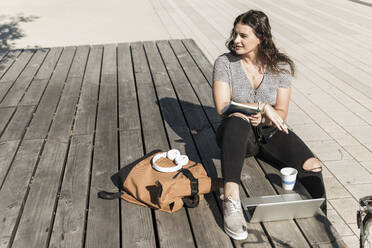 Nachdenkliche junge Frau, die mit Laptop und Rucksack auf der Strandpromenade in der Stadt sitzt und wegschaut - UUF20750