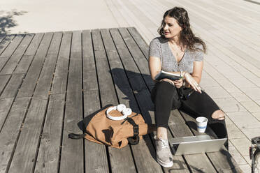 Thoughtful young woman looking away while sitting with laptop and backpack on boardwalk in city - UUF20750