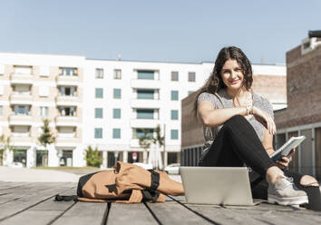 Smiling young woman with laptop and backpack sitting on boardwalk in city - UUF20751