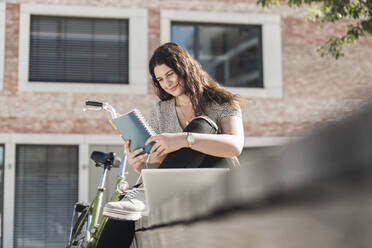 Lächelnde junge Frau liest ein Buch, während sie an einem Gebäude in der Stadt sitzt - UUF20752