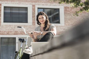 Smiling young woman holding smart phone while sitting against building in city - UUF20755