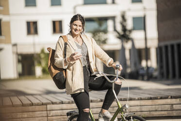 Smiling female commuter holding smart phone riding bicycle in city during sunny day - UUF20760