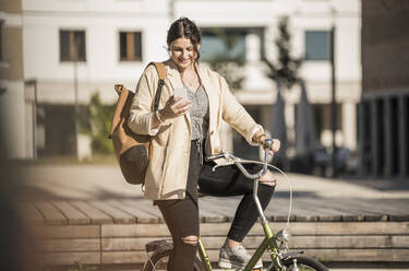 Studentin, die auf dem Fahrrad sitzend in der Stadt an einem sonnigen Tag ein Mobiltelefon benutzt - UUF20761