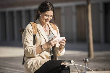 Junge Frau, die ihr Smartphone benutzt, während sie auf einem Fahrrad in der Stadt bei Sonnenschein sitzt - UUF20763