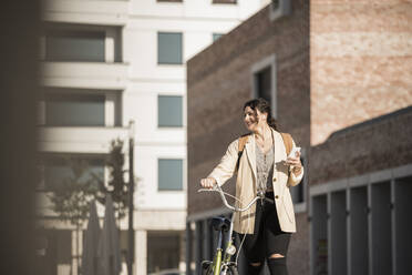 Smiling female student looking away while walking with bicycle against buildings - UUF20765