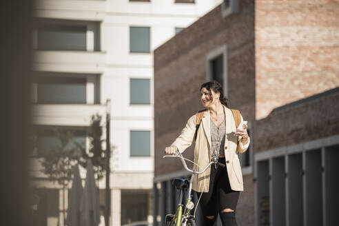 Lächelnde Studentin, die wegschaut, während sie mit dem Fahrrad gegen ein Gebäude fährt - UUF20765