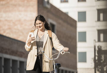 Smiling female student using smart phone while walking with bicycle against buildings in city - UUF20766