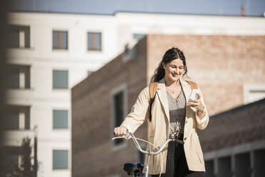 Smiling female commuter using mobile phone while walking with bicycle against buildings in city - UUF20767