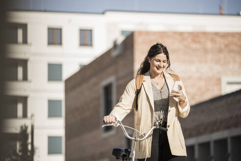 Lächelnder weiblicher Pendler, der ein Mobiltelefon benutzt, während er mit dem Fahrrad gegen Gebäude in der Stadt läuft - UUF20767