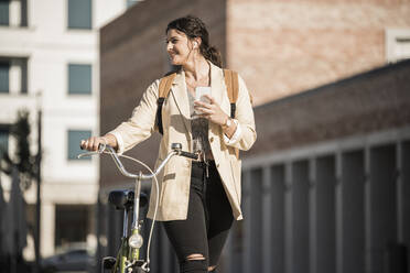 Lächelnde Studentin, die wegschaut, während sie mit dem Fahrrad gegen Gebäude in der Stadt fährt - UUF20769