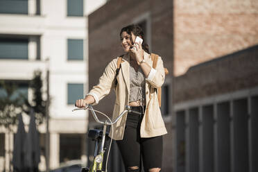 Lächelnde junge Frau, die mit dem Handy telefoniert, während sie mit dem Fahrrad in der Stadt spazieren geht - UUF20772