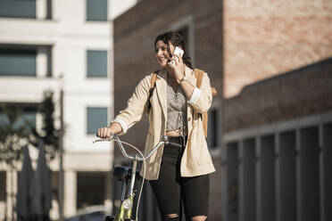 Smiling young woman talking over mobile phone while walking with bicycle in city - UUF20772