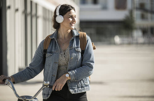 Lächelnde Frau hört Musik über Kopfhörer, während sie mit dem Fahrrad in der Stadt spazieren geht - UUF20775