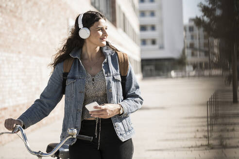 Junge Frau hört Musik und schaut weg, während sie mit dem Fahrrad in der Stadt steht - UUF20778