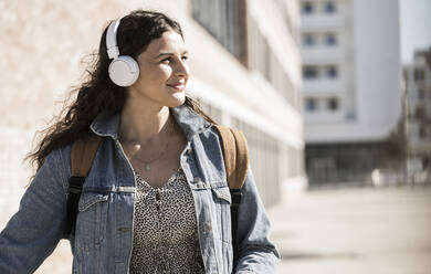 Junge Frau hört Musik über Kopfhörer und schaut weg, während sie in der Stadt steht - UUF20780