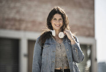 Smiling young woman with headphones standing in city on sunny day - UUF20782