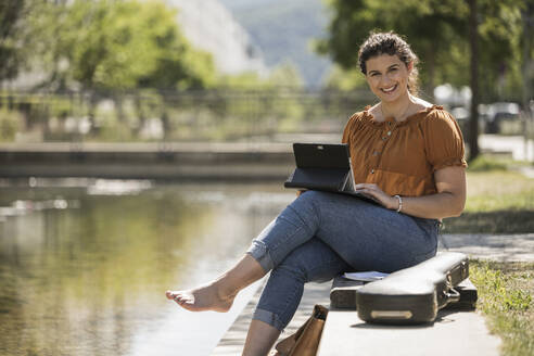Lächelnde junge Frau, die einen Laptop benutzt, während sie an einem sonnigen Tag am Teich im Park sitzt - UUF20785