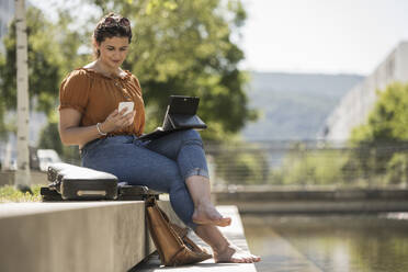 Frau mit Laptop und Smartphone im Park sitzend an einem sonnigen Tag - UUF20786