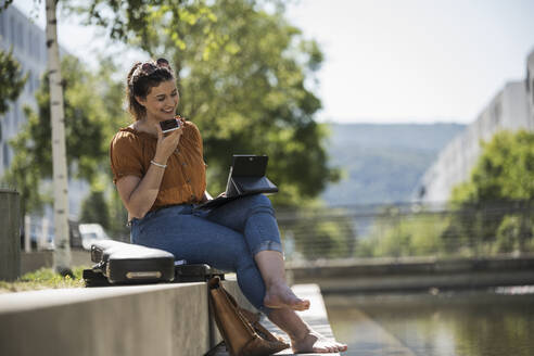 Lächelnde Frau mit Laptop, die über ihr Smartphone spricht, während sie an einem sonnigen Tag im Park sitzt - UUF20787