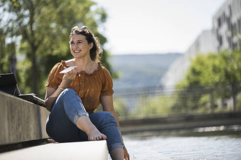 Lächelnde junge Frau, die im Park sitzend über ihr Mobiltelefon spricht - UUF20792