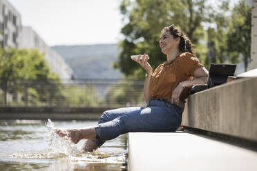 Cheerful woman with legs in pond talking over smart phone while sitting at park - UUF20794