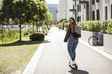 Lächelnde Frau fährt elektrischen Roller auf der Straße in der Stadt während des sonnigen Tages - UUF20799