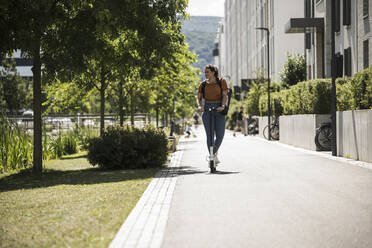 Young woman riding electric push scooter on road in city during sunny day - UUF20800