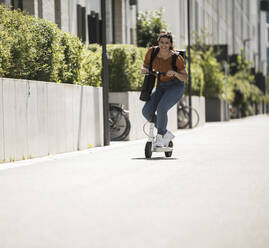 Lächelnde Frau auf elektrischem Roller auf der Straße an einem sonnigen Tag - UUF20801