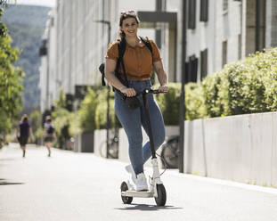 Lächelnde junge Frau auf elektrischem Roller auf der Straße an einem sonnigen Tag - UUF20802