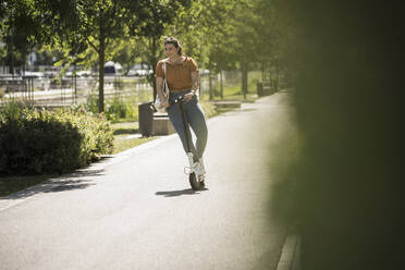 Young woman riding electric push scooter on road against trees during sunny day - UUF20803