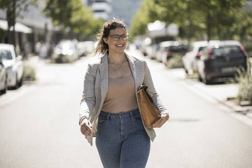 Lächelnde junge Frau mit Tasche auf der Straße an einem sonnigen Tag - UUF20804