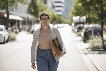 Lächelnde junge Frau, die eine Tasche trägt, während sie an einem sonnigen Tag auf der Straße geht - UUF20805