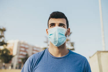 Close-up of thoughtful young man wearing mask against clear sky in city - MIMFF00108