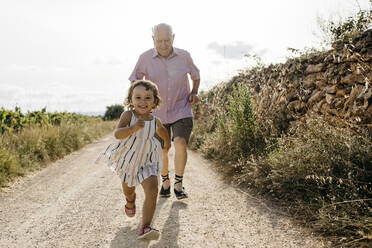 Großvater läuft hinter spielerischen Enkelin auf unbefestigten Weg gegen Himmel - JRFF04650
