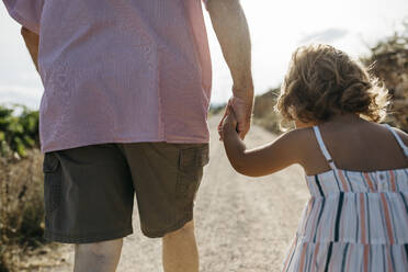 Close-up of grandfather holding granddaughter's hand while walking on dirt road - JRFF04652