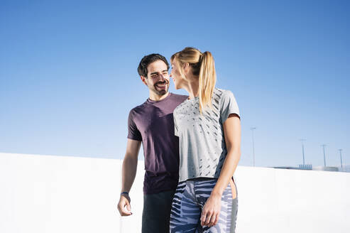 Smiling couple talking while standing against clear blue sky in city during sunny day - JCMF01094