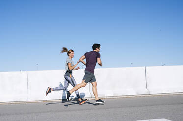 Couple running on road against clear blue sky in city during sunny day - JCMF01095