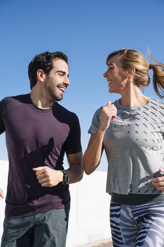 Smiling couple looking at each other while running against clear blue sky on sunny day stock photo