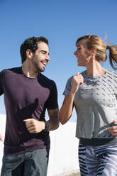 Smiling couple looking at each other while running against clear blue sky on sunny day - JCMF01096