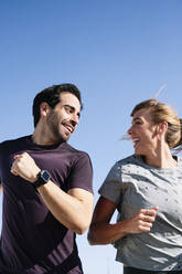 Smiling couple looking at each other while running against clear blue sky - JCMF01097