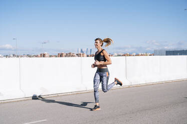 Woman running on road against blue sky in city during sunny day - JCMF01100