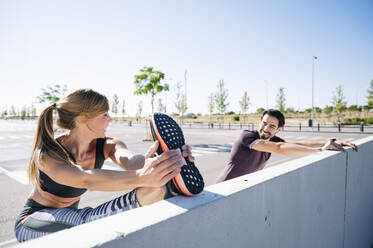 Smiling couple stretching on retaining wall against clear sky during sunny day - JCMF01104