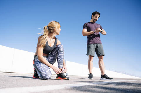 man checking time while woman tying shoelace on road against clear blue sky in city stock photo