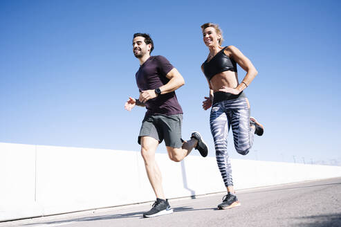 Smiling couple running on street against clear blue sky in city during summer - JCMF01107