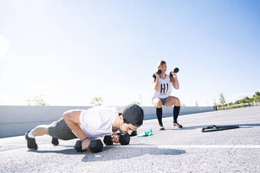Couple with dumbbells exercising on road against clear sky during sunny day - JCMF01120