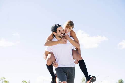 Happy man piggybacking woman while walking against sky during sunny day - JCMF01124