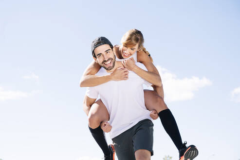 Cheerful man piggybacking woman while walking against sky during sunny day - JCMF01125