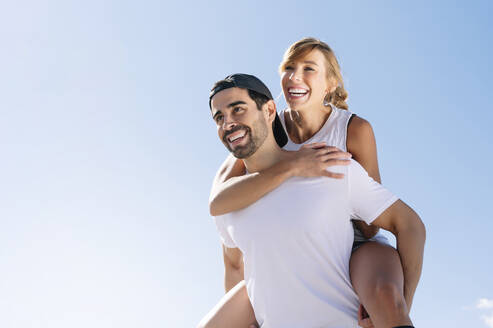 Mid adult man piggybacking cheerful woman against clear sky during sunny day - JCMF01126